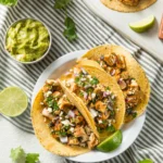 Chicken street tacos with fresh toppings on a rustic table, accompanied by salsa and guacamole.